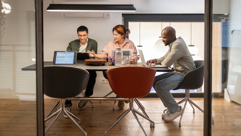 A diverse team collaborates around a table in a modern coworking office. They are engaged in discussion, using laptops and documents to facilitate their meeting.