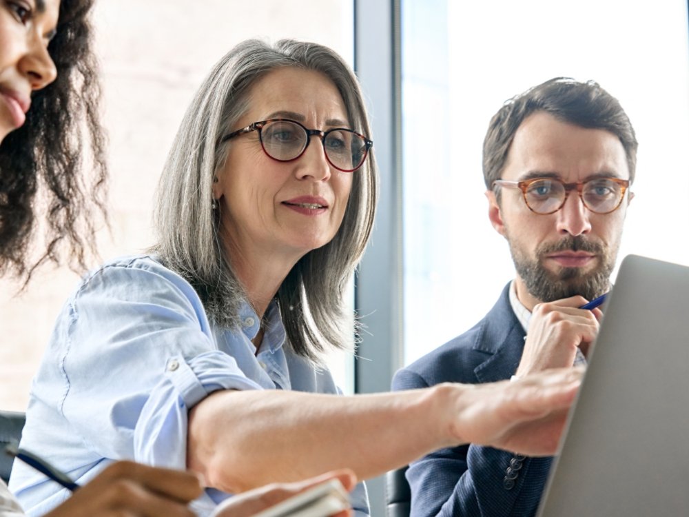 Mature older ceo businesswoman mentor in glasses negotiating growth business plan with diverse executive managers at boardroom meeting table using laptop. Multicultural team work together in office.