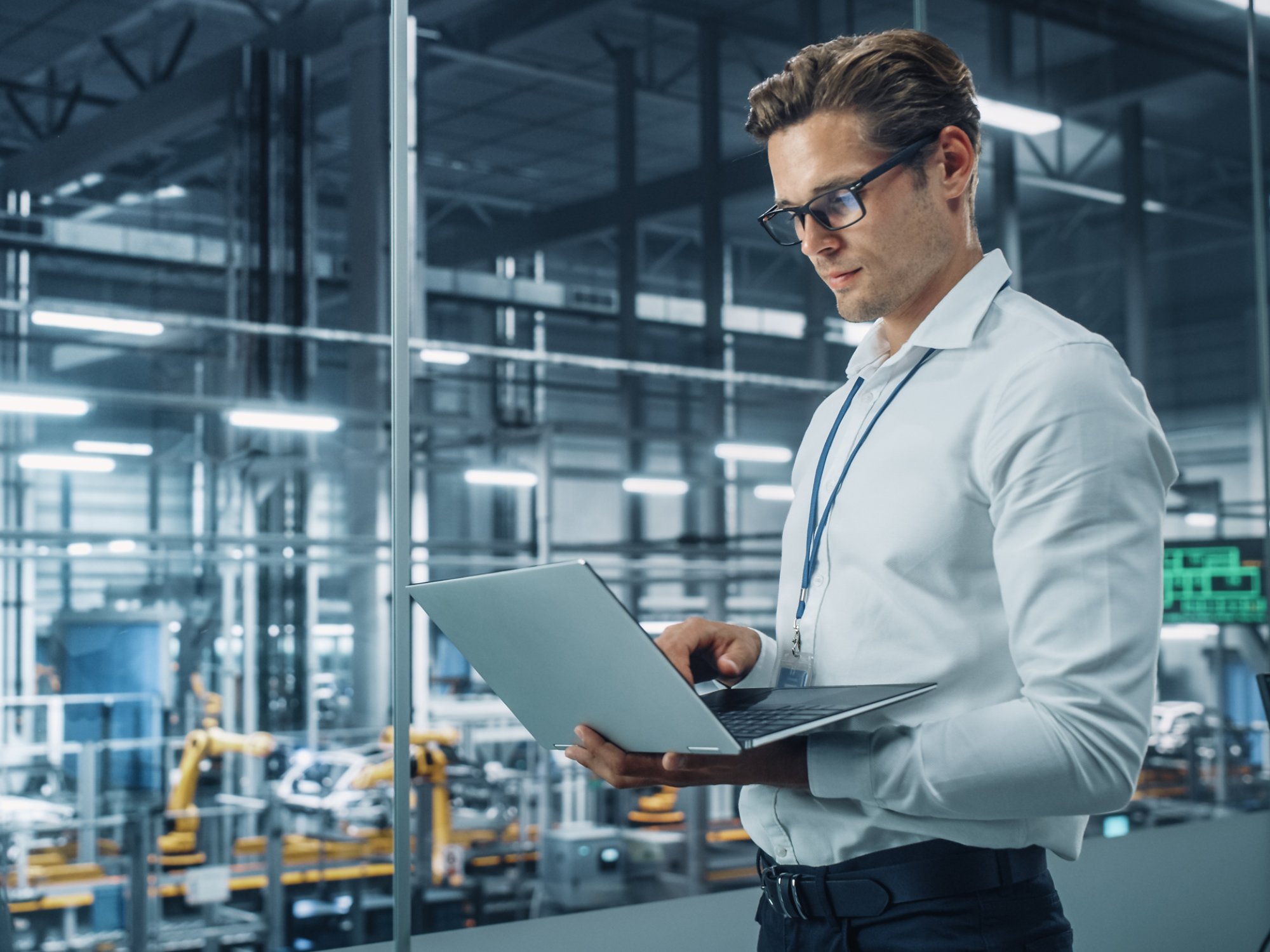 Handsome Engineer in Glasses and White Shirt Using Laptop Computer and Looking Out of the Office at a Car Assembly Plant. Industrial Specialist Working on Vehicle Design in Technological Facility.