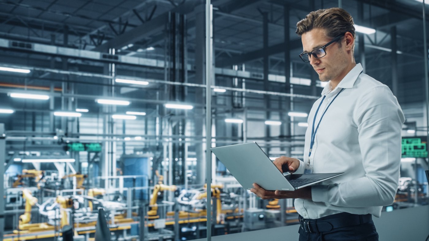 Handsome Engineer in Glasses and White Shirt Using Laptop Computer and Looking Out of the Office at a Car Assembly Plant. Industrial Specialist Working on Vehicle Design in Technological Facility.