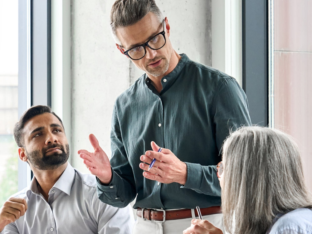 Mature businessman leader mentor talking to diverse colleagues team listening to caucasian ceo. Multicultural professionals project managers group negotiating in boardroom at meeting. Vertical.