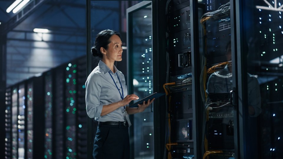 A female IT specialist stands in a modern data center holding a digital tablet. She inspects a tall server rack filled with illuminated hardware and network cables. Long rows of server cabinets extend into the background, creating a high-tech environment. Cool blue lighting and reflections on the glass doors emphasize the futuristic, professional atmosphere.