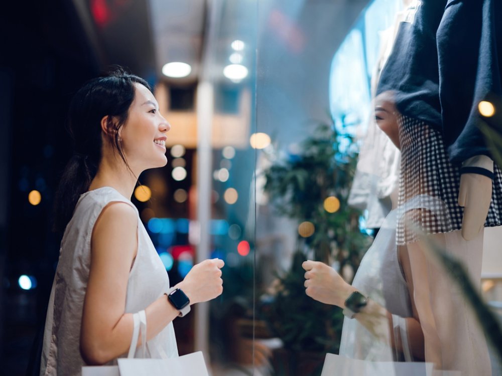 Smiling young Asian woman carrying a paper shopping bag, standing outside a boutique looking at shop window in the evening in city