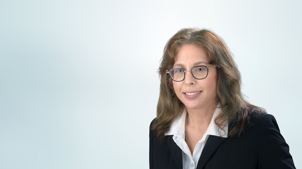 A professional woman wearing glasses and a dark blazer over a white shirt poses for a studio headshot. She is positioned on the right side of the frame against a soft light blue gradient background. Her expression is friendly and approachable, suitable for corporate or business use. The clean, minimal backdrop emphasizes her face and formal attire.