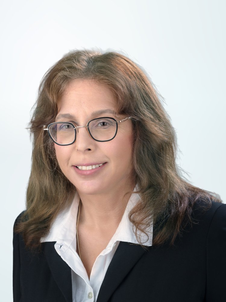 A professional woman wearing glasses and a dark blazer over a white shirt poses for a studio headshot. She is positioned on the right side of the frame against a soft light blue gradient background. Her expression is friendly and approachable, suitable for corporate or business use. The clean, minimal backdrop emphasizes her face and formal attire.