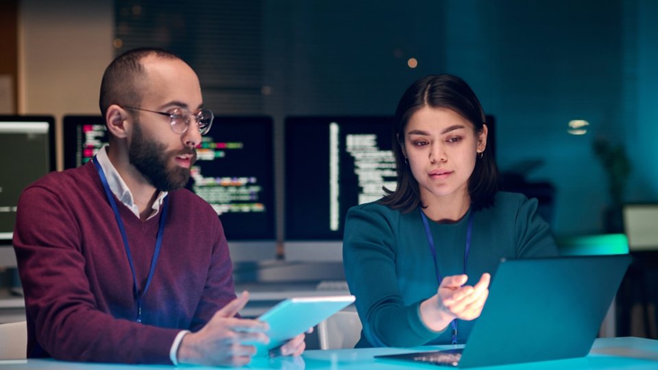 Portrait of young woman as female IT security expert using laptop with male colleague in blue neon light
