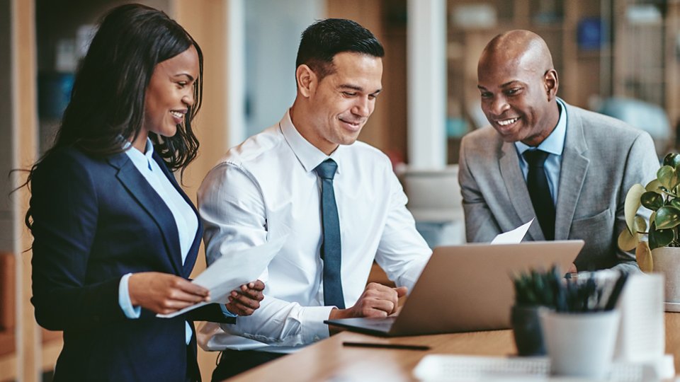 Smiling group of diverse businesspeople going over paperwork together and working on a laptop at a table in an office