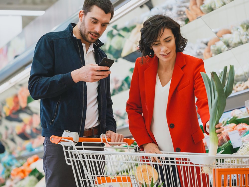 At the Supermarket: Happy Couple Does Shopping, Choosing Fruits and Vegetables in the Fresh Produce Section. Man Uses Smartphone and Pushes Shopping Cart, Woman Chooses Products.