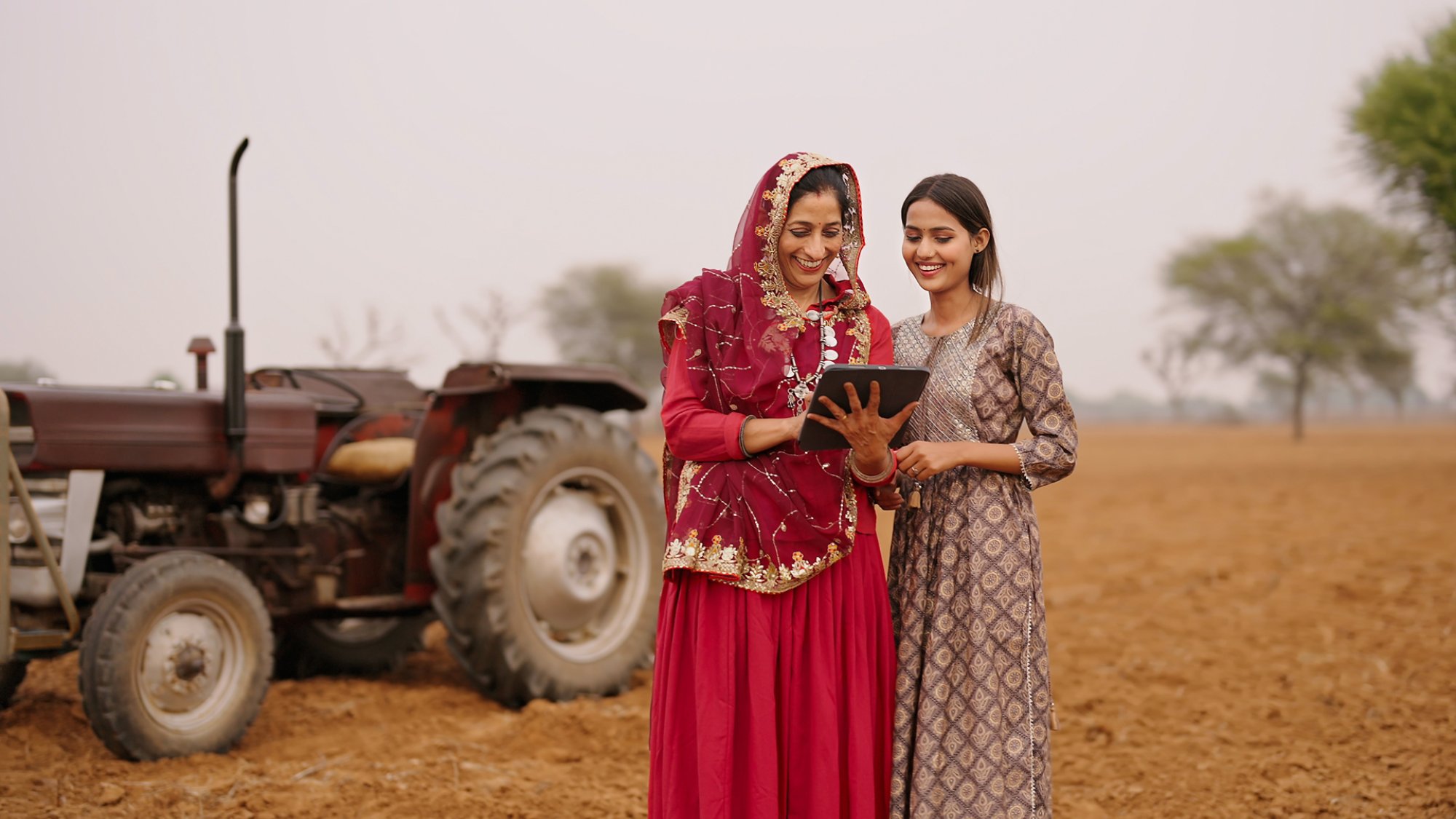 Indian asian villager farmer young adult woman lady wear red saree smiling outdoor hold tab device browse website data happy teen age girl teach mom talking enjoy day time outside desi life