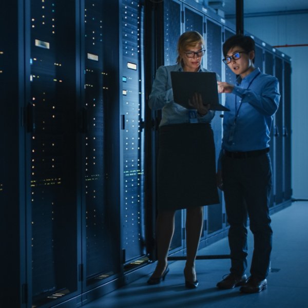 In the Modern Data Center: Engineer and IT Specialist Work with Server Racks, Talking, Doing System Maintenance Check with Laptop Computer. Concept of Diagnostics of the Database, Cybersecurity Check