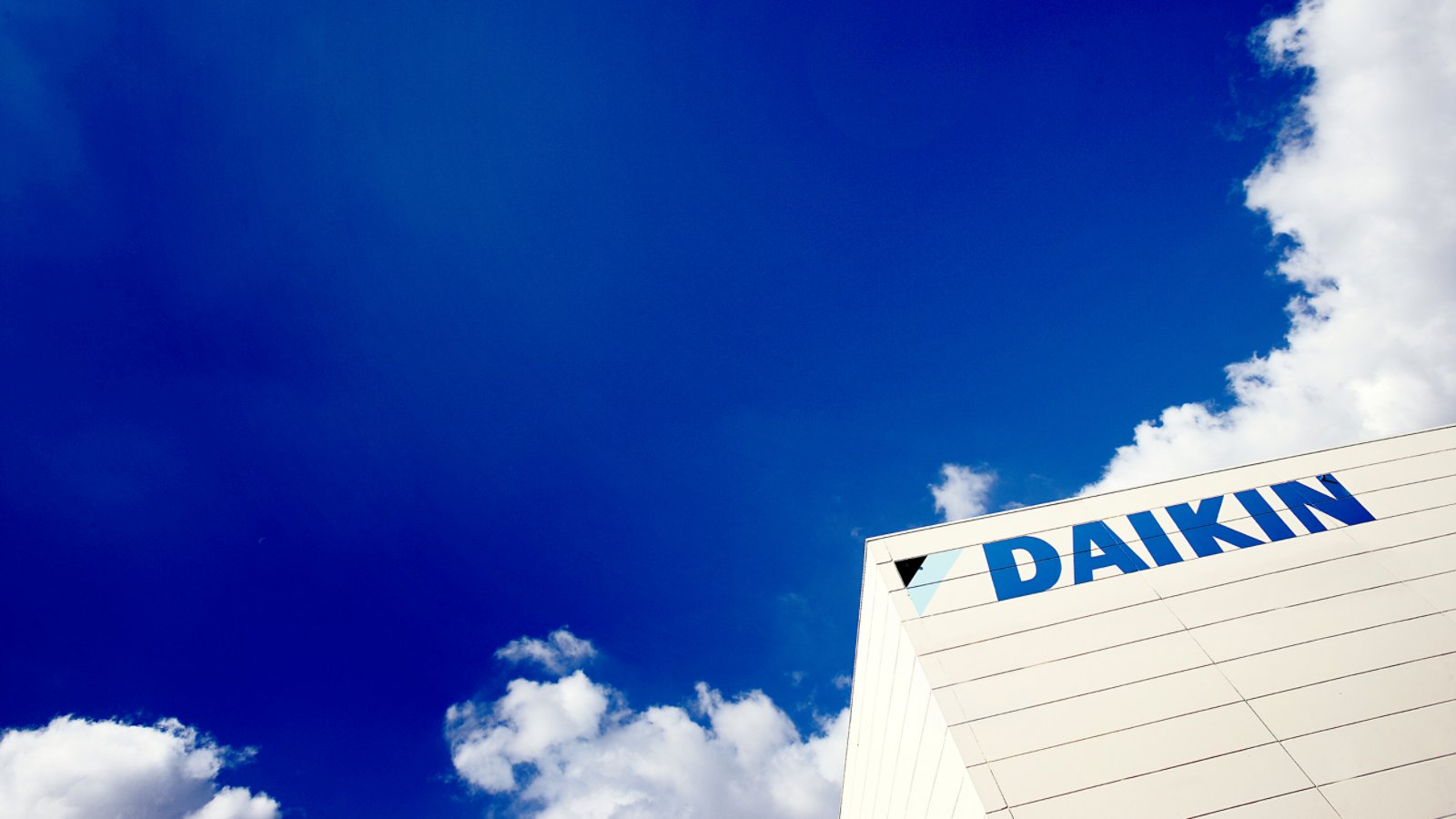 A modern industrial building corner displays the Daikin name prominently against a bright blue sky. The white facade is clean and minimal, emphasizing the bold blue text and triangular symbol. Fluffy white clouds frame the structure, creating a sense of openness and good weather. The composition highlights the contrast between the geometric architecture and the expansive sky.