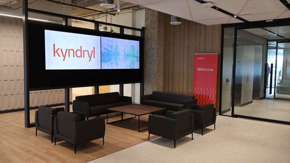 A contemporary office lobby features a seating area with black armchairs and a wooden coffee table on a light floor. Two large digital screens display the Kyndryl name and abstract graphics on the left wall. To the right, a red vertical banner reads “Kyndryl Welcome” near a glass-walled meeting room. The space has an open, minimalist design with exposed ceiling elements and neutral tones.