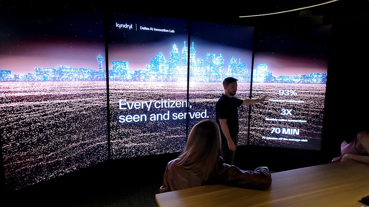 A man stands in front of a large multi-panel digital display, pointing to data visualizations and text. The screen shows a stylized city skyline above a glowing field of data points, with phrases like "Every citizen, seen and served" and numerical metrics including 93% and 70 MIN. Several people sit at a wooden table watching the presentation in a dark, modern room. The scene suggests a technology or innovation lab environment focused on data-driven urban services.