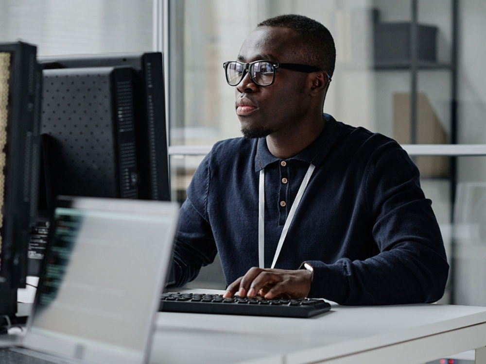 African American young developer in eyeglasses concentrating on his online work on computer sitting at workplace