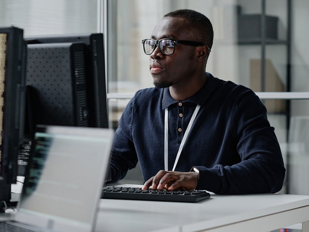 African American young developer in eyeglasses concentrating on his online work on computer sitting at workplace