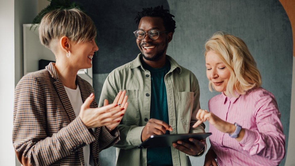 Three smiling colleagues are discussing a project, using a digital tablet and exchanging ideas in a modern office environment, showcasing teamwork and collaboration