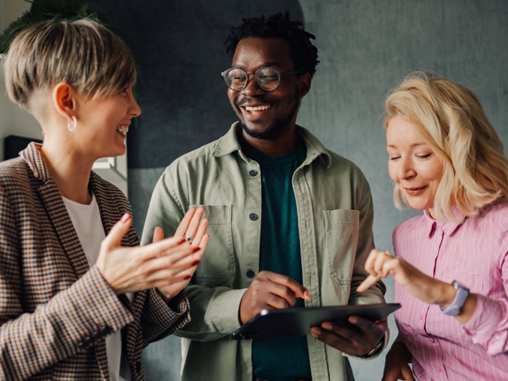 Three smiling colleagues are discussing a project, using a digital tablet and exchanging ideas in a modern office environment, showcasing teamwork and collaboration