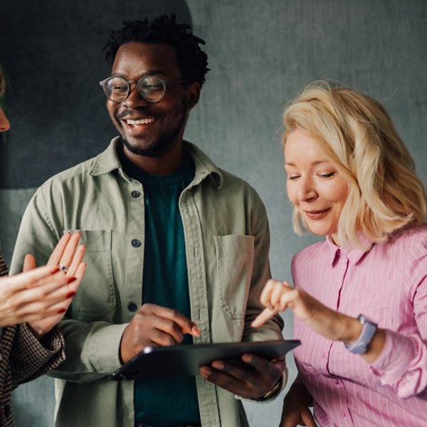 Three smiling colleagues are discussing a project, using a digital tablet and exchanging ideas in a modern office environment, showcasing teamwork and collaboration