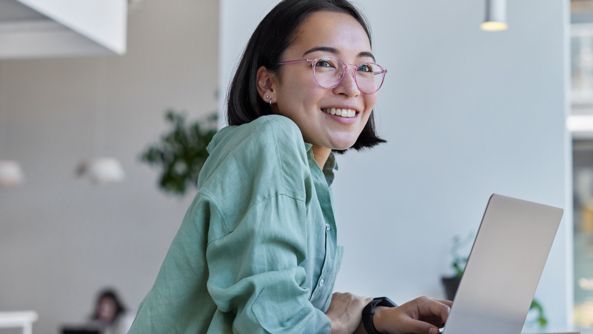 Pretty cheerful Asian woman in eyeglasses and casual clothes browses laptop computer connected to 4g internet updates software uses modern technologies poses in cafeteria looks gladfully away