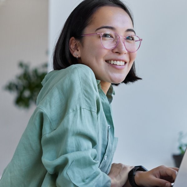 Pretty cheerful Asian woman in eyeglasses and casual clothes browses laptop computer connected to 4g internet updates software uses modern technologies poses in cafeteria looks gladfully away
