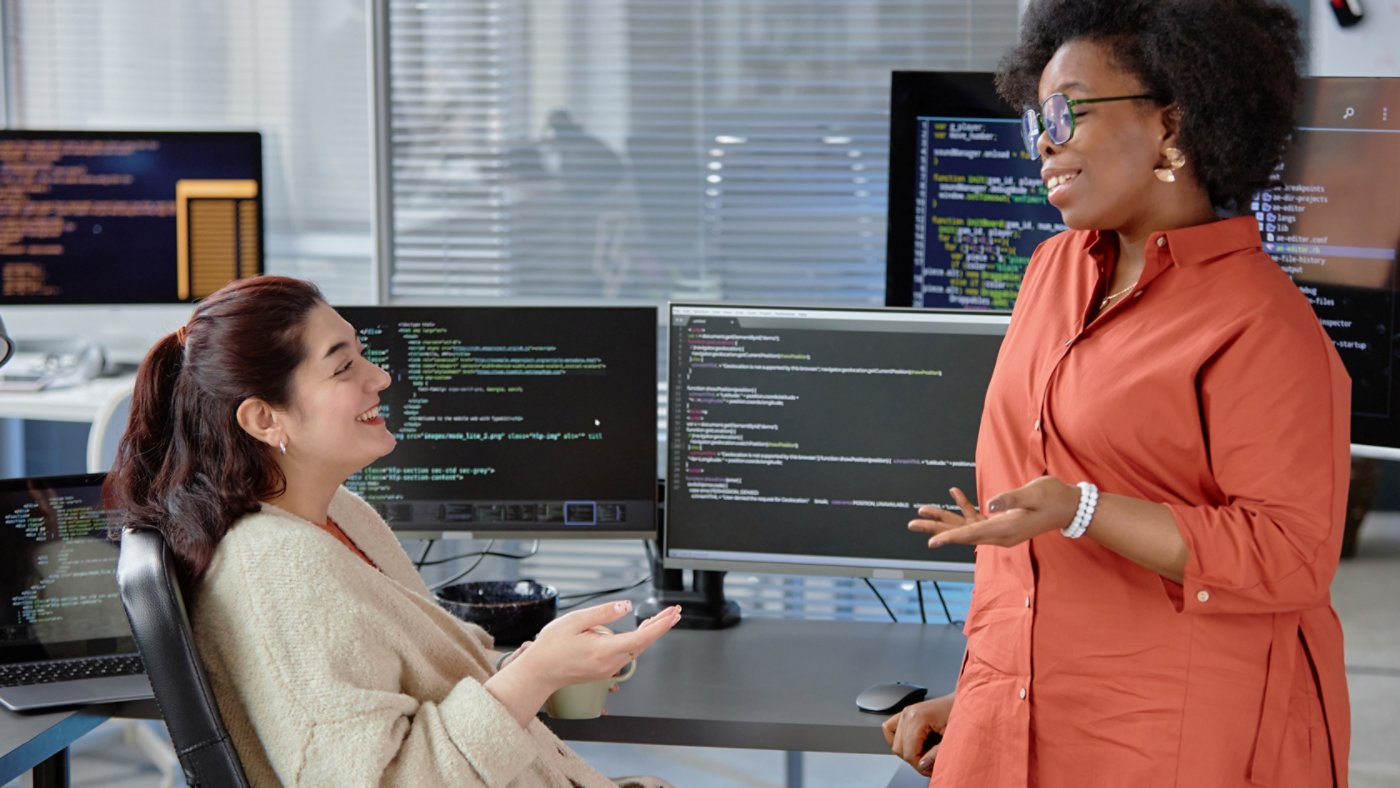 Side view shot of diverse group of two larger-bodied cheerful software developers smiling, while casually chatting and discussing program code at desk with computer screens in cutting-edge office