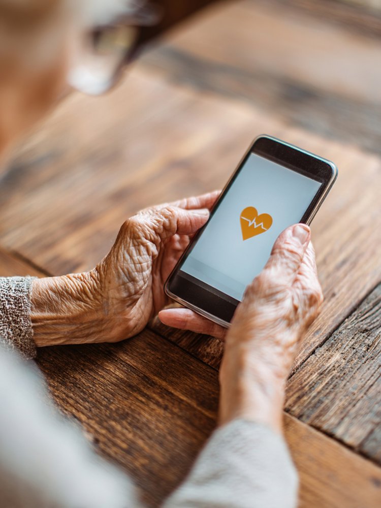 A close-up of an elderly woman holding a modern smartphone with a heart-shaped icon on it, resting on a warm wooden table. 