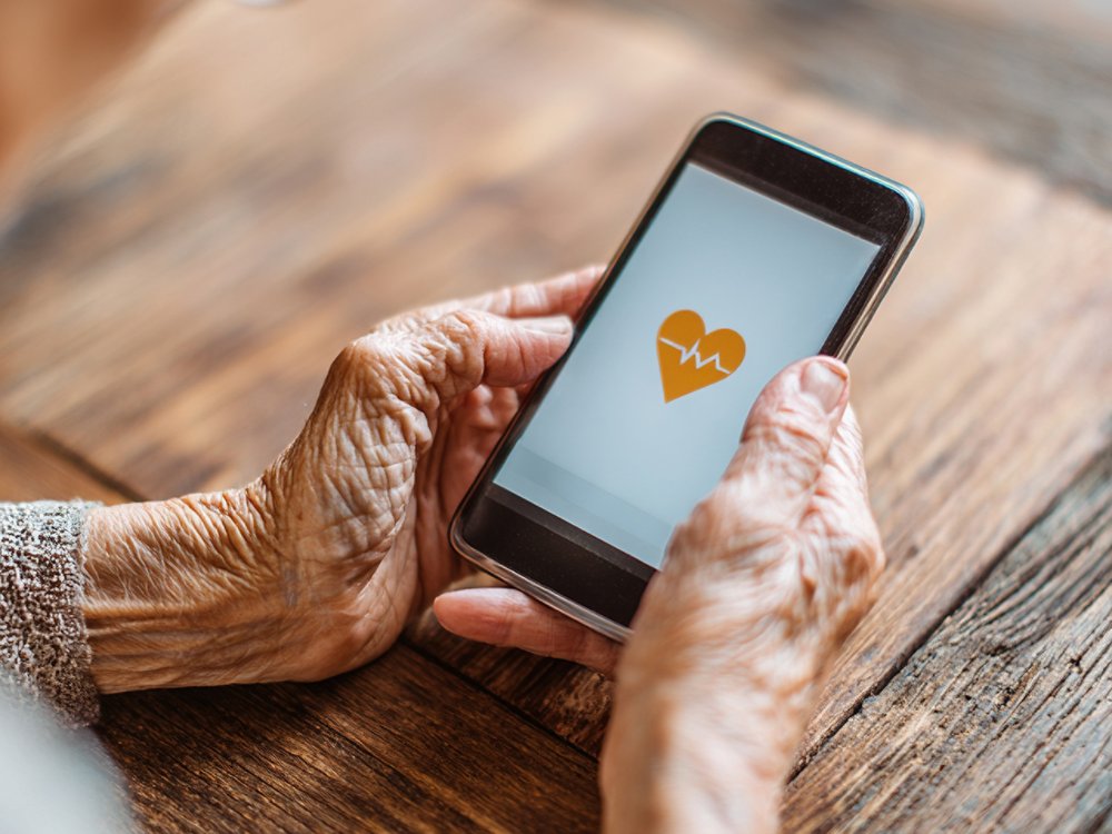 A close-up of an elderly woman holding a modern smartphone with a heart-shaped icon on it, resting on a warm wooden table. 