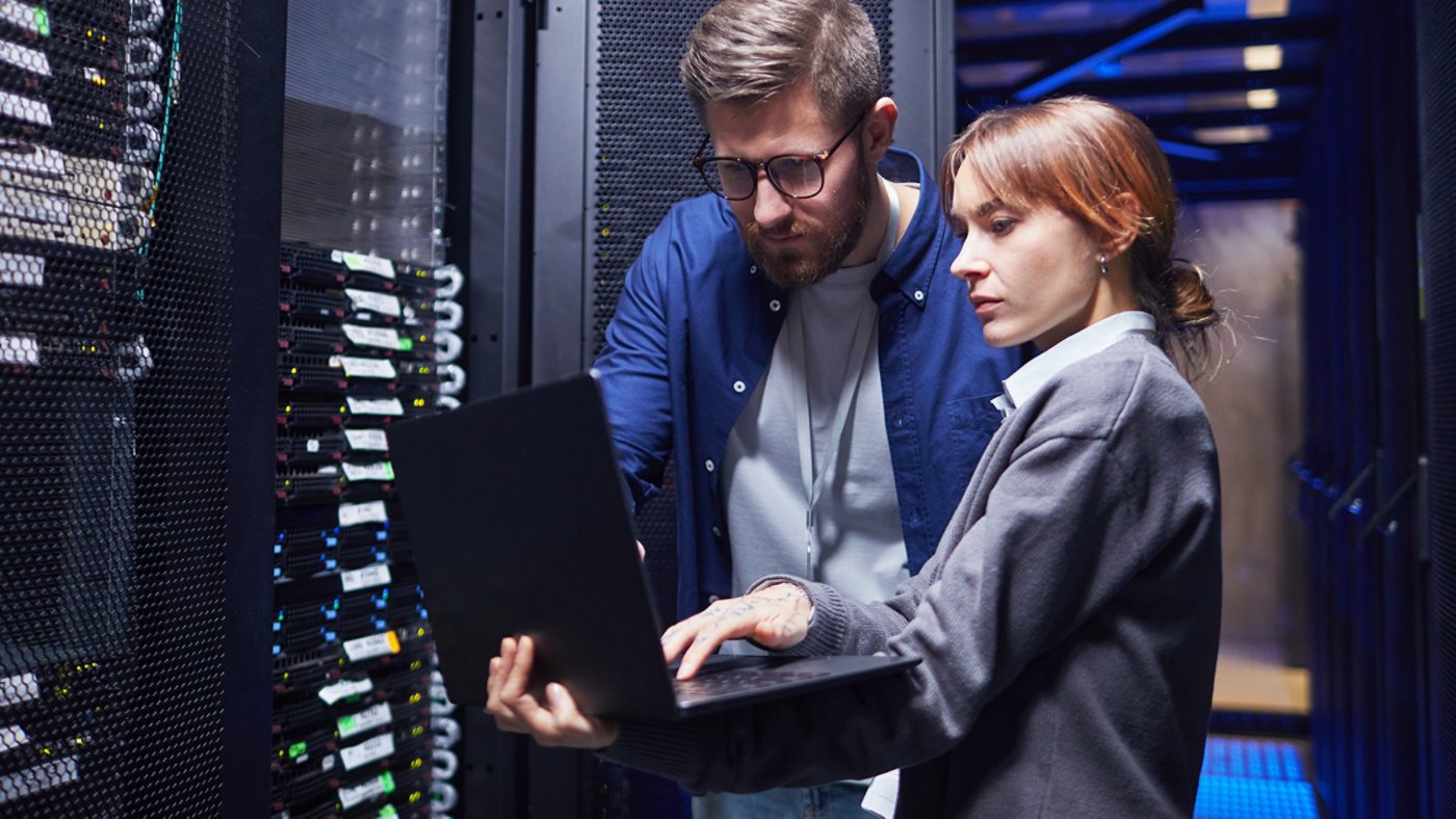 Two IT professionals are examining server connections in a data center, focusing on laptop screen while discussing necessary steps for troubleshooting issues