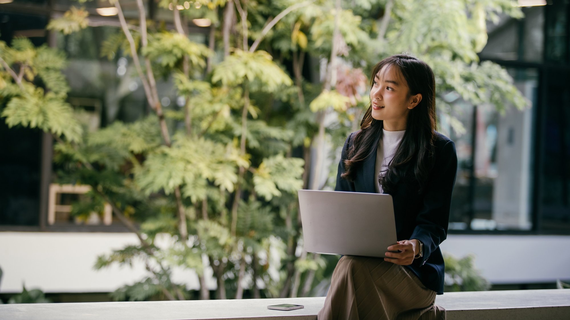 Young businesswoman sits with her laptop in a modern office space adorned with lush greenery, looking thoughtfully towards upcoming projects and opportunities