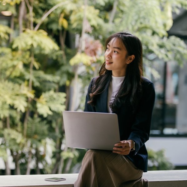 Young businesswoman sits with her laptop in a modern office space adorned with lush greenery, looking thoughtfully towards upcoming projects and opportunities