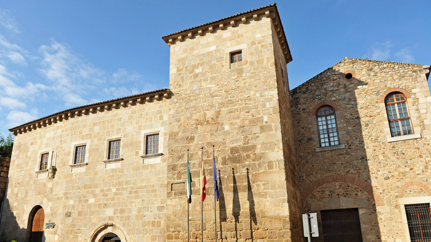 A large, historic stone building is shown under a bright blue sky with scattered clouds. The structure features small rectangular windows, a prominent tower, and several flags displayed at the entrance. The sunlight casts clear shadows, highlighting the texture of the stone walls. No visible text or numbers are present in the image.