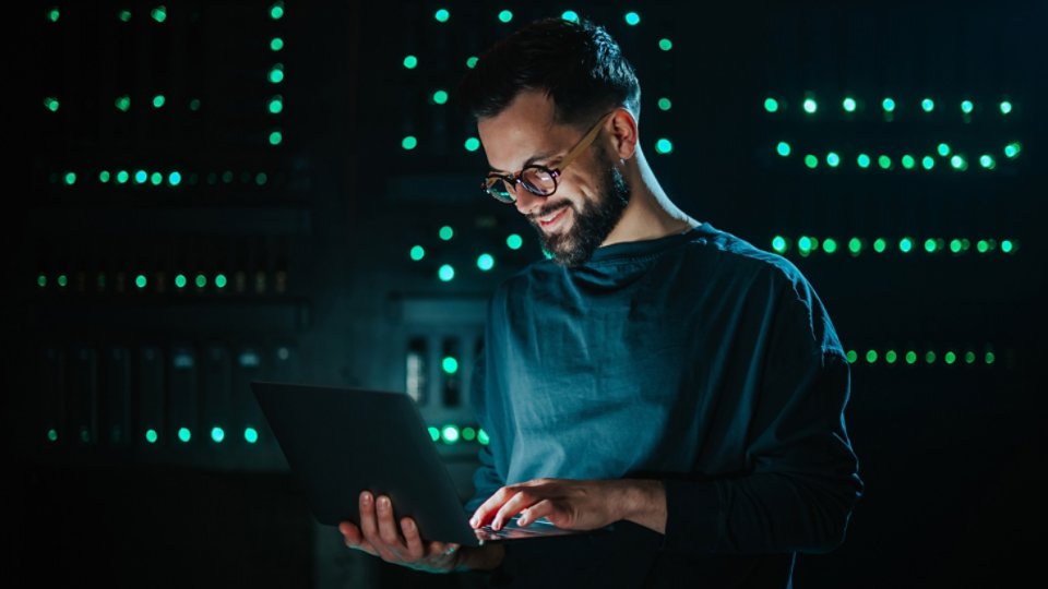 Young smiling it professional using laptop in server room, performing network engineering and cybersecurity tasks for data management