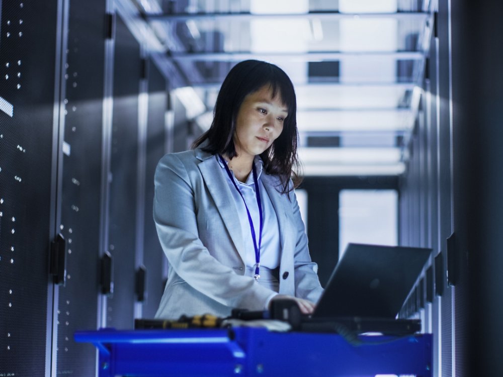 Asian Female IT Engineer Working on a Laptop on Tool Cart, She Scans Hard Drives.  She's in a Big Data Center Full of Rack Servers.
