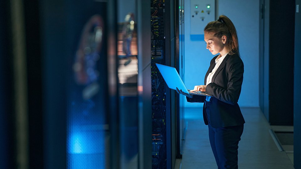Woman IT specialist in elegant suit working on notebook computer in data center next to server racks