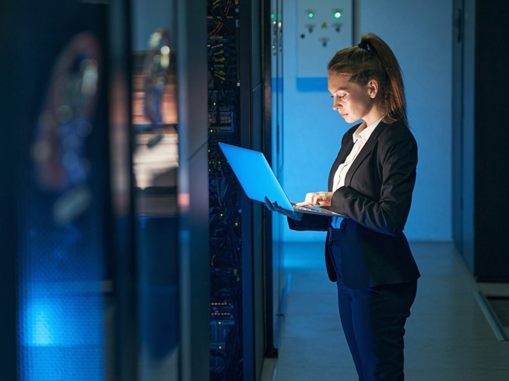 Woman IT specialist in elegant suit working on notebook computer in data center next to server racks