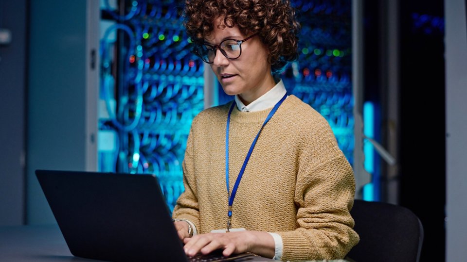 Young engineer woman sitting at table and typing data on laptop, she working over new system online in data center