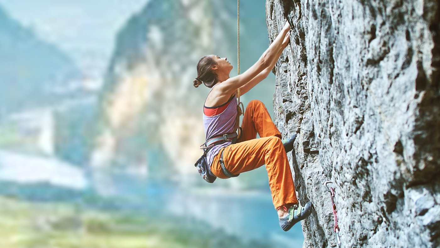 side view of young slim woman rock climber in bright orange pants climbing on the cliff against a high mountains. girl climbs on a vertical flat rocky wall and making hard move. Copy space