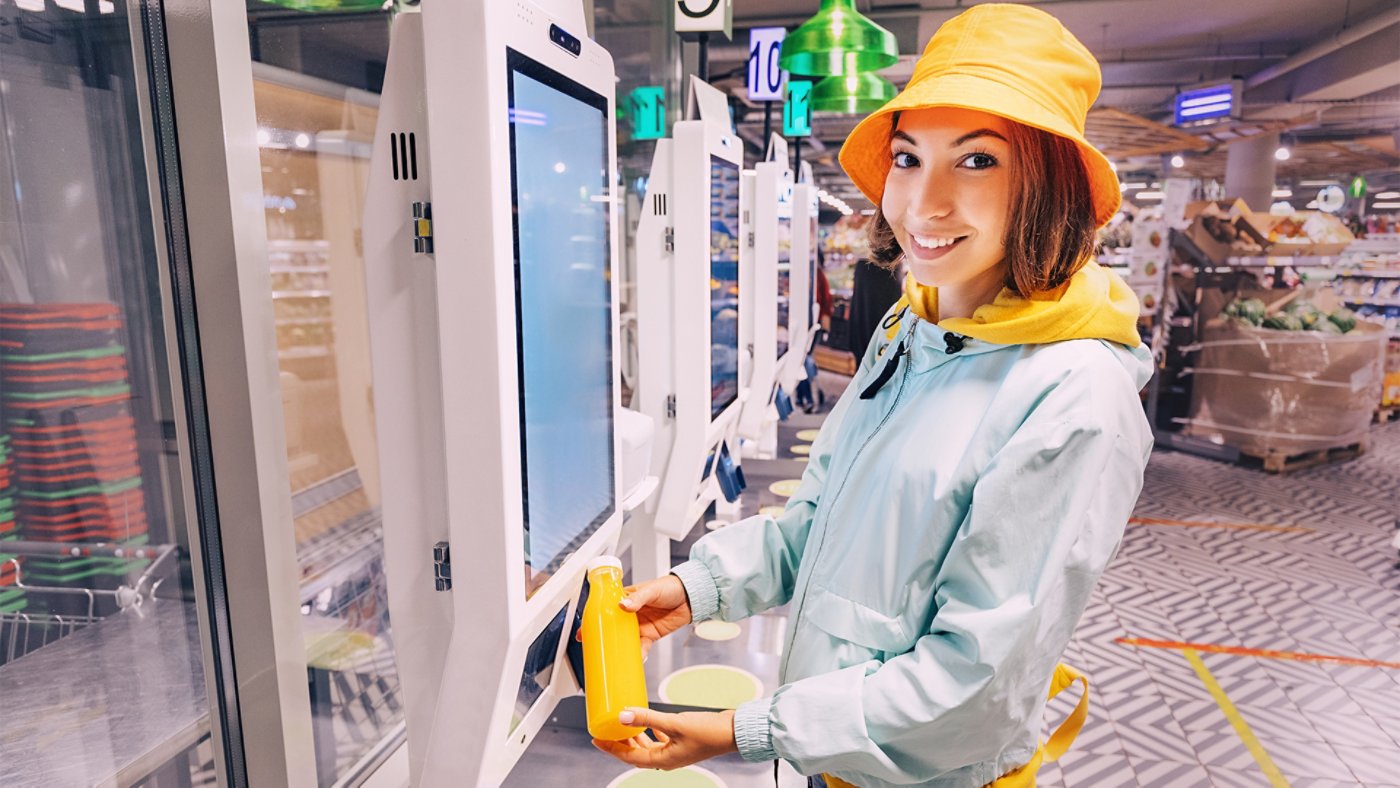 Happy young customer woman buys and pays for a bottle of orange juice at the checkout of a self-service vending machine in a modern supermarket, to avoid waiting in line