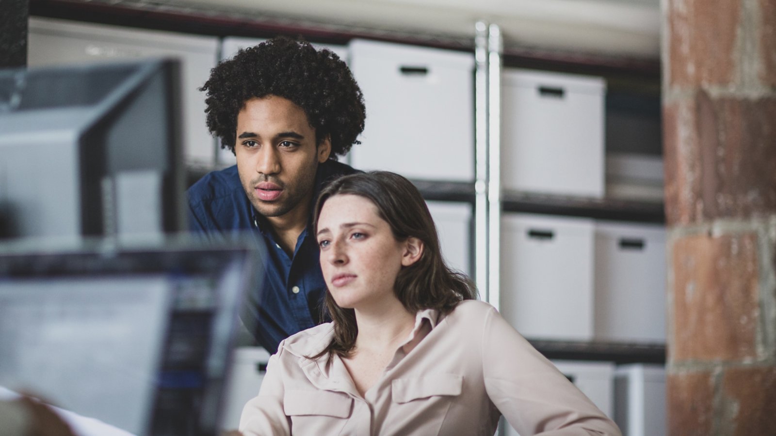Two individuals are working together in a modern office setting with visible storage boxes in the background. The environment suggests a collaborative workspace with a focus on organization and productivity. A computer monitor is partially visible, indicating a digital work context.