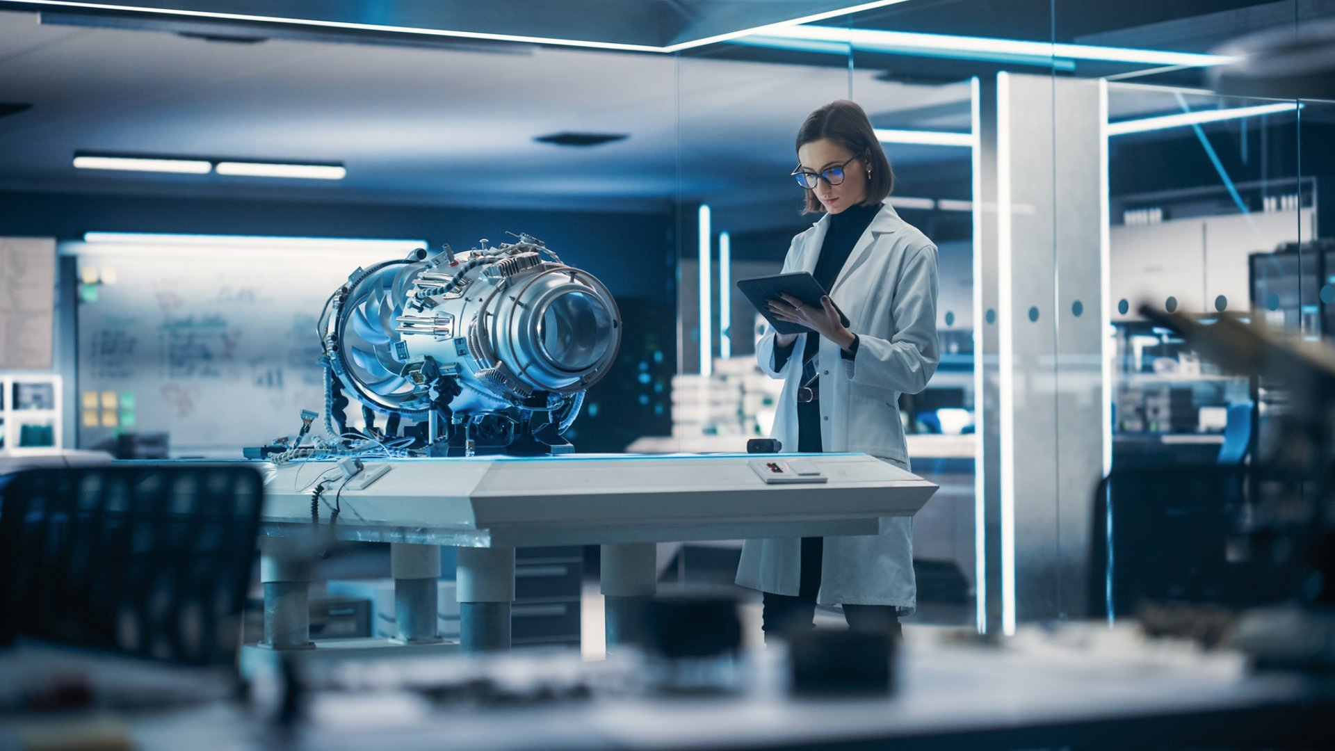 A female scientist in a white lab coat stands in a high-tech laboratory, examining data on a digital tablet. In front of her, a large cylindrical mechanical device rests on a central workbench, illuminated by cool blue lighting. The lab is filled with glass walls, metallic surfaces, and glowing screens, creating a futuristic atmosphere. The scene suggests advanced research, engineering, or experimental technology development.