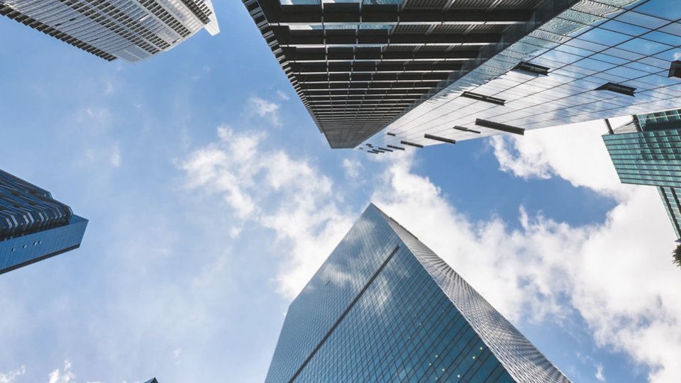A cluster of tall, modern skyscrapers viewed from below, set against a bright blue sky with scattered clouds. The buildings feature reflective glass facades and sleek architectural designs. The image emphasizes urban development and contemporary cityscapes.