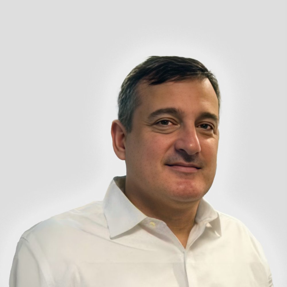The image shows an adult man wearing a white button-up shirt against a plain light gray background. His face is intentionally blurred, emphasizing anonymity and privacy. The neutral backdrop and simple clothing suggest a professional or corporate context. The overall composition is clean and minimalistic, suitable for use as a generic profile or placeholder portrait.