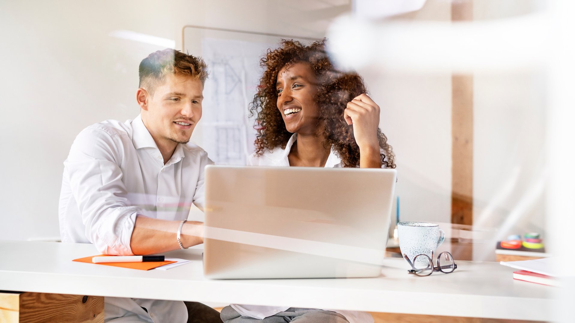 Smiling colleagues discussing over laptop on desk in office seen through window