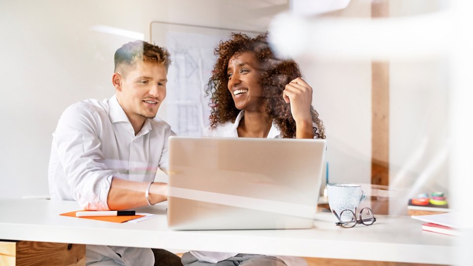 Smiling colleagues discussing over laptop on desk in office seen through window