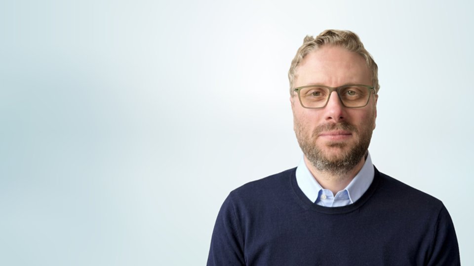 A man wearing glasses is shown in a front-facing portrait against a light, gradient background. He has short, light-colored hair and a trimmed beard. The man is dressed in a dark sweater over a collared shirt, giving a professional appearance. The composition is minimalistic, with the subject positioned to the right and ample negative space on the left.