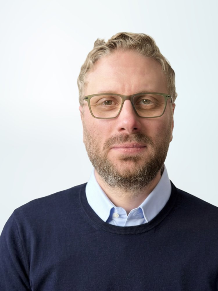 A man wearing glasses is shown in a front-facing portrait against a light, gradient background. He has short, light-colored hair and a trimmed beard. The man is dressed in a dark sweater over a collared shirt, giving a professional appearance. The composition is minimalistic, with the subject positioned to the right and ample negative space on the left.