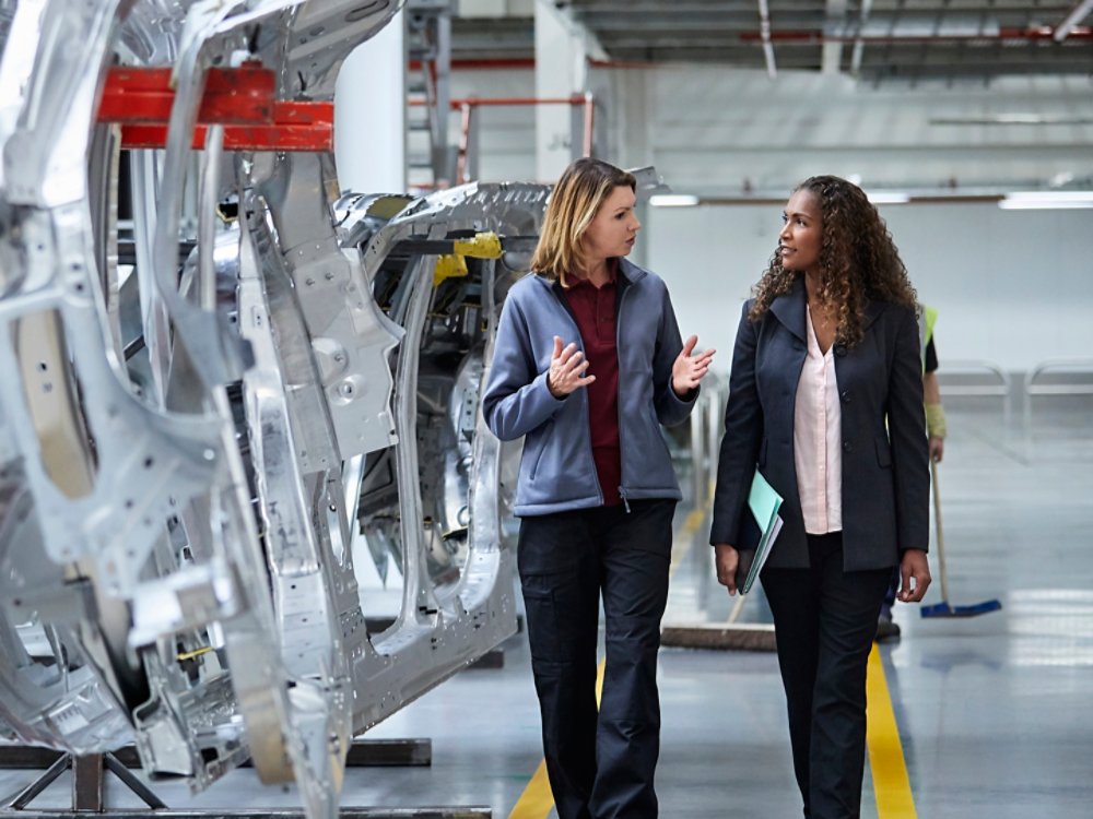 Female engineers discussing by car chassis. Full length of professionals are walking on aisle in factory. Colleagues are in automobile industry.