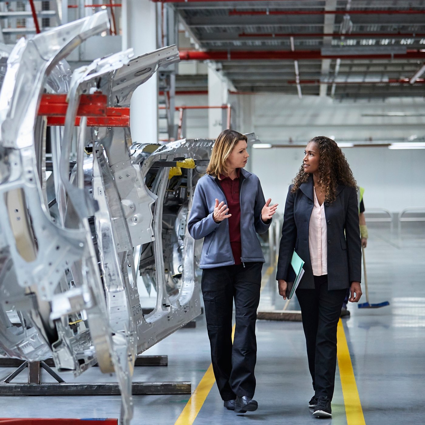 Female engineers discussing by car chassis. Full length of professionals are walking on aisle in factory. Colleagues are in automobile industry.