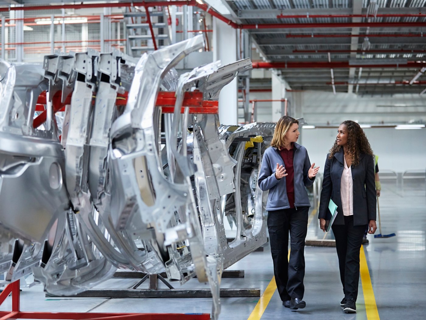 Female engineers discussing by car chassis. Full length of professionals are walking on aisle in factory. Colleagues are in automobile industry.