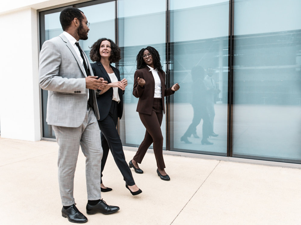 Multiethnic business team walking near office building. Man and women wearing formal suits, going past outdoor wall, talking to each other. Walking colleagues concept